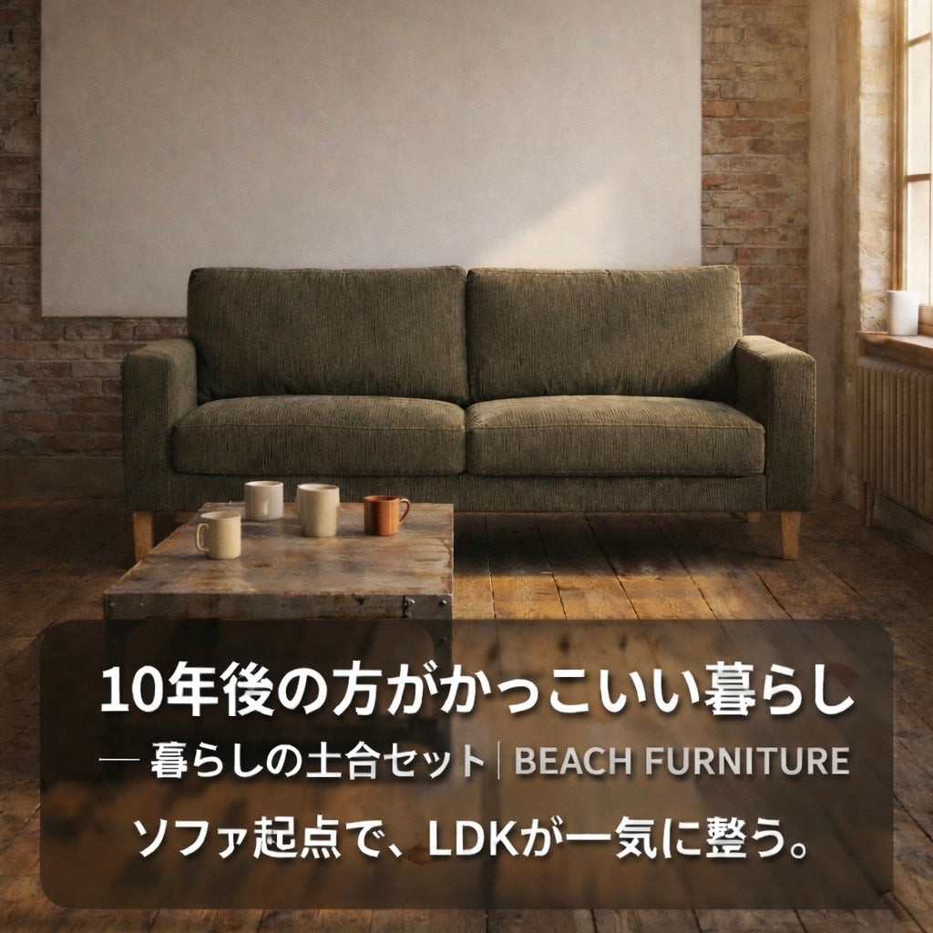 レンガ壁と大きな白いボードのある部屋に、オリーブグリーンの2人掛けソファと木脚、手前にマグカップを置いたローテーブル。右側の窓から柔らかな光が入り、「10年後の方がかっこいい暮らし/暮らしの土台セット|BEACH FURNITURE/ソファ起点でLDKが一気に整う。」のテキストが重なる。
