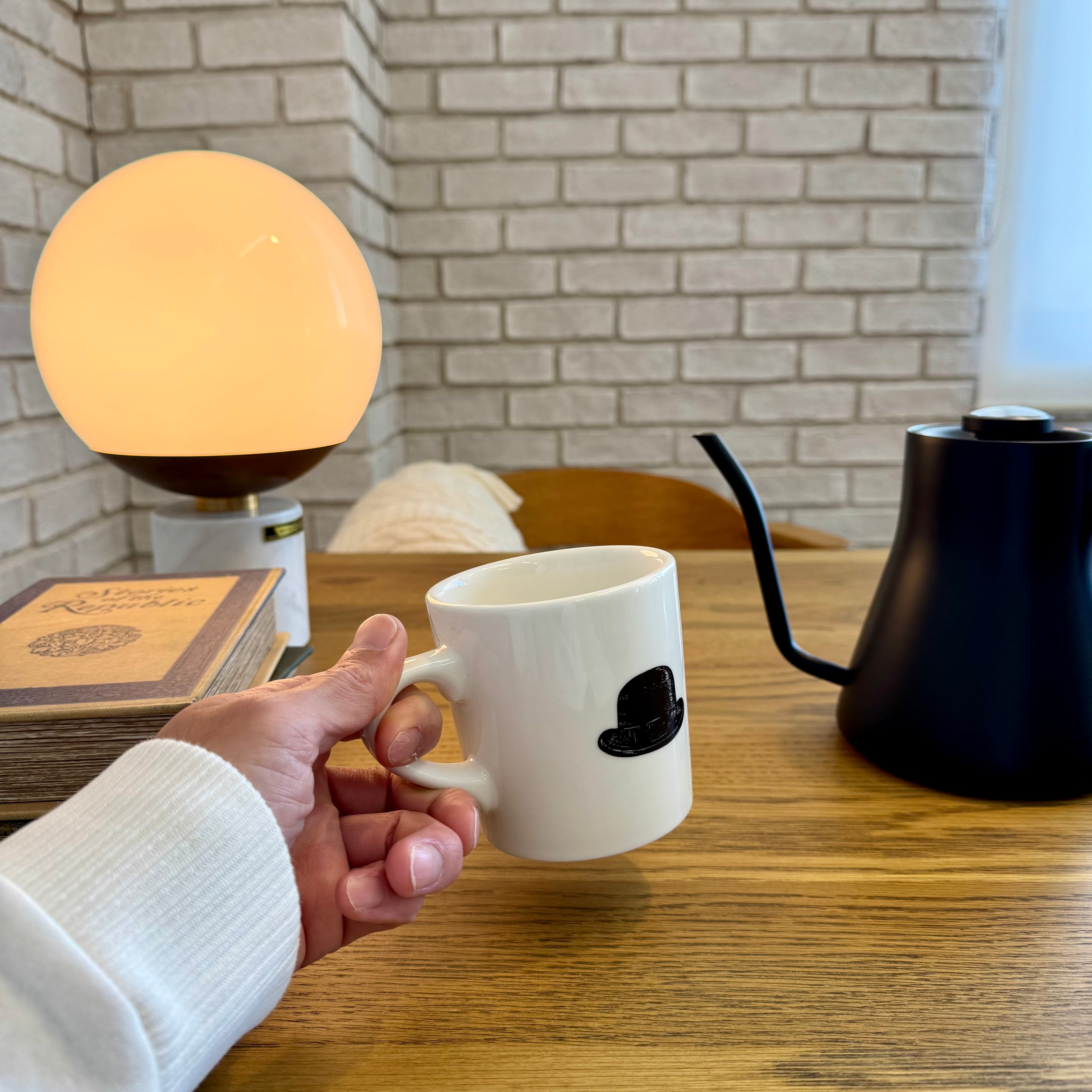 マグを手に持つ手元のアップ。無垢材のBYRONダイニングテーブル天板の木目と、丸いテーブルランプの柔らかな灯りが写る。